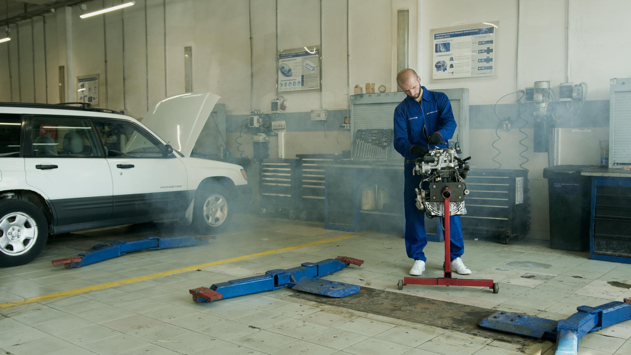 Mechanic in blue overalls working on a car engine in an auto repair shop with a white car.